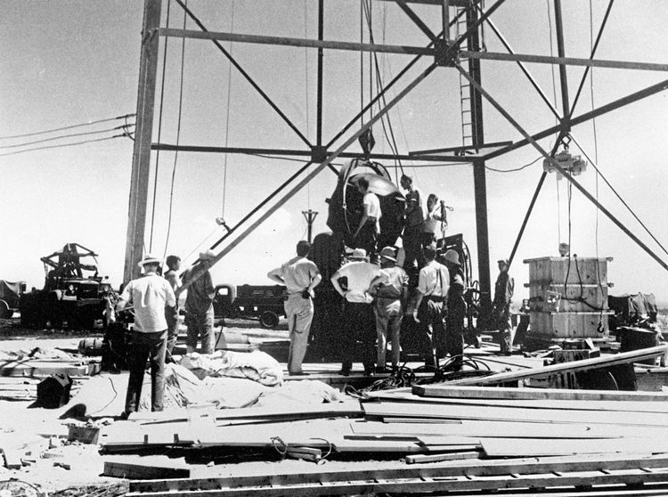 Mehrere Personen stehen um den Atombombenprototyp, der an einer Seilwinde hängt, um ihn auf den Turm zu ziehen.
