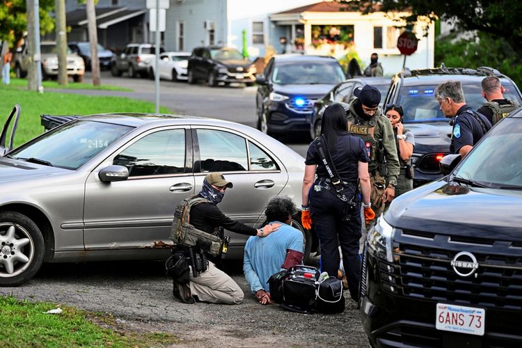 Ein ICE-Agent kniet neben einer festgenommenen Person, während Sanitäter des Danbury Emergency Medical Services (EMS) medizinische Hilfe leisten. Im Hintergrund sind mehrere Polizeifahrzeuge mit eingeschaltetem Blaulicht vor einem Wohnhaus in Danbury, Connecticut, zu sehen.