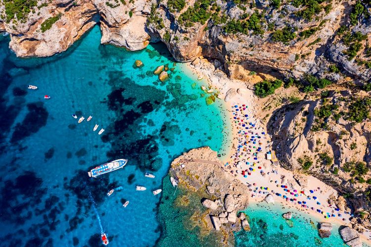 Luftaufnahme eines Strandes mit türkisfarbenem Wasser, umgeben von felsigen Klippen. Mehrere bunte Sonnenschirme verteilen sich auf dem Sandstrand, während Boote im klaren Meer vor Anker liegen. Die Umgebung ist grün und felsig. Italien, Sardinien.