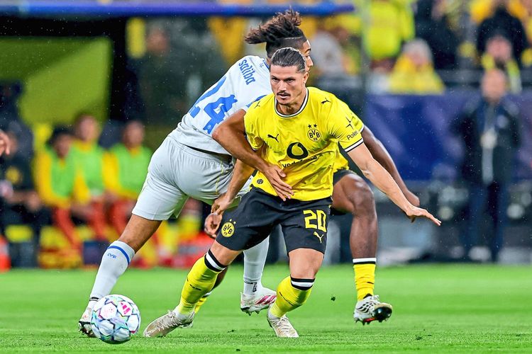 Ein Fußballspieler von Borussia Dortmund (im gelben Trikot) kämpft um den Ball mit einem Spieler des Villarreal CF (im weißen Trikot) während eines Champions-League-Spiels im Signal Iduna Park. Im Hintergrund sind Fans und Teamkollegen zu sehen.