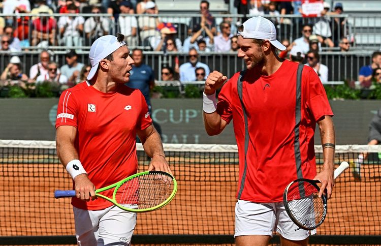 Zwei Tennisspieler in roten Trikots, Lucas Miedler und Neil Oberleitner, feiern auf einem Sandplatz ihren Sieg im Davis-Cup-Doppelspiel. Im Hintergrund sitzen Zuschauer auf Tribünen.