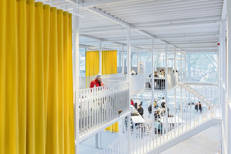 Das Gebäude von innen: Decken, Böden, Handläufe und Geländer - alles ist weiß, dazwischen hängen zwei gelbe Vorhänge; auf zwei Etagen sind Studierende beim Lernen zu sehen; dahinter eine große Glasfront, die einen Blick ins Grüne freigibt