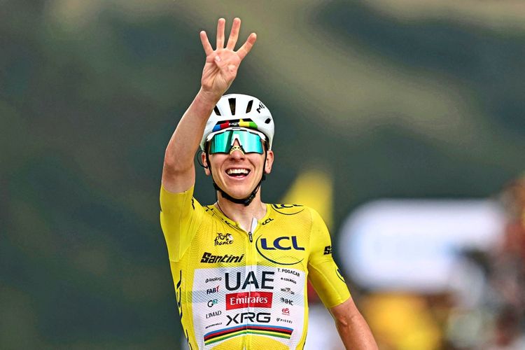 Ein Radfahrer im gelben Trikot des Gesamtführenden streckt vier Finger in die Luft und lächelt, während er das Ziel einer Etappe der Tour de France erreicht. Im Hintergrund sind unscharfe Landschaft und Zuschauer zu sehen.