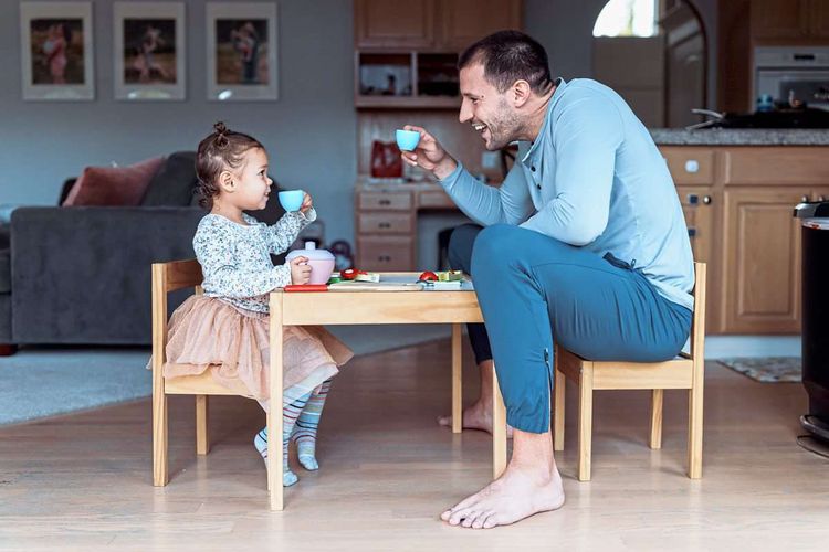 Ein Papa sitzt mit seiner Tochter auf einem kleinen Tisch. Beide halten eine kleine Teetasse in der Hand und lachen.