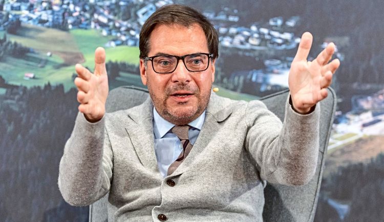 Ein Mann mit Brille, grauem Wollblazer und Krawatte gestikuliert in einer Gesprächssituation. Im Hintergrund sind unscharf eine Landschaft und Gebäude zu sehen.