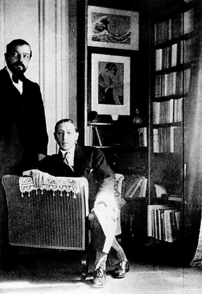 Schwarz-weißes Foto von Claude Debussy (stehend) und Igor Strawinsky (sitzend) in einer Wohnung mit Bücherregal und ein Kunstdruck von Hokusai an der Wand.