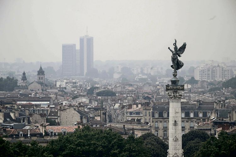 Eine Statue auf einer Säule ist vor dem Hintergrund der Stadt Bordeaux zu sehen, die in dichten Rauch gehüllt ist. Der Rauch stammt von nahegelegenen Waldbränden, die die Skyline und die umliegende Landschaft am 26. Juli 2026 stark beeinflussen.