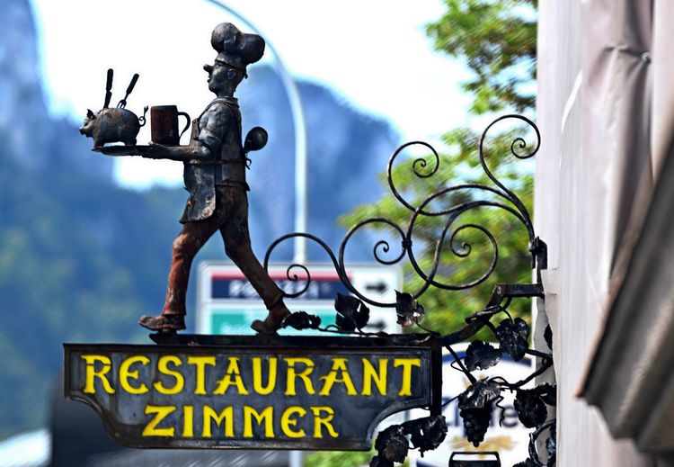 Ein kunstvoll gestaltetes Schild mit der Aufschrift „Restaurant-Zimmer“ in gelber Schrift. Oben ist eine Metallfigur eines Kochs mit Tablett und Utensilien dargestellt. Hintergrund mit unscharfer Vegetation und Verkehrszeichen.