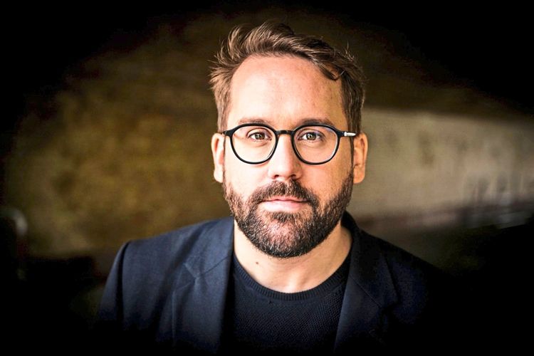 Ein Mann mit Brille und Bart, der einen dunklen Blazer und ein schwarzes Oberteil trägt, blickt direkt in die Kamera. Der Hintergrund ist unscharf und in dunklen Tönen gehalten.