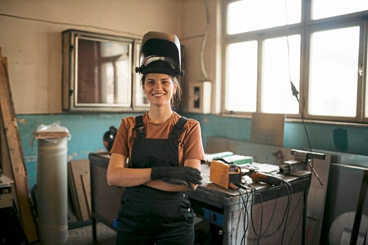 Junge Frau mit hochgeklappter Schweißermaske steht in einer Werkstatt und lächelt in die Kamera
