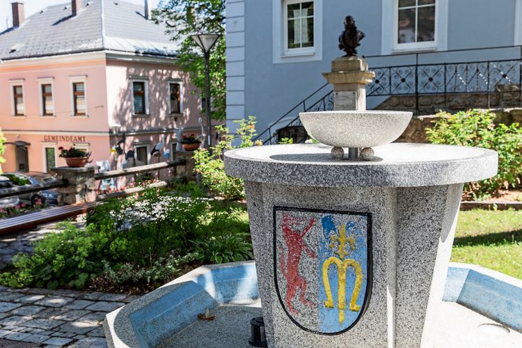 Ein steinerner Brunnen mit Wappen verziert, das einen roten Löwen und ein gelbes Symbol mit blauem Hintergrund zeigt. Im Hintergrund ist ein rosafarbenes Gebäude mit der Aufschrift 