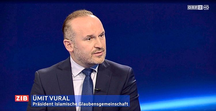 Ein Mann mit Anzug und Krawatte spricht in einer Studioaufnahme vor einem dunkelblauen Hintergrund. Unten eingeblendet steht: 