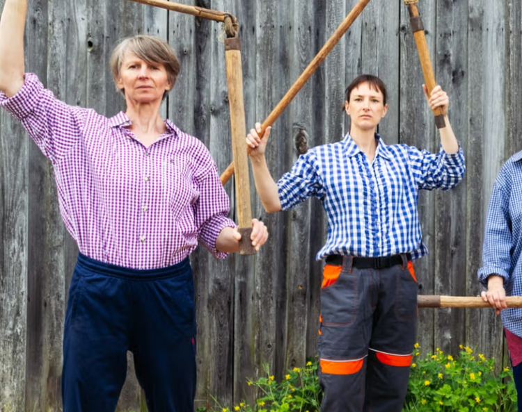 Drei Personen in karierten Hemden stehen vor einer Holzwand und halten traditionelle Sensen oder Dreschwerkzeuge hoch.
