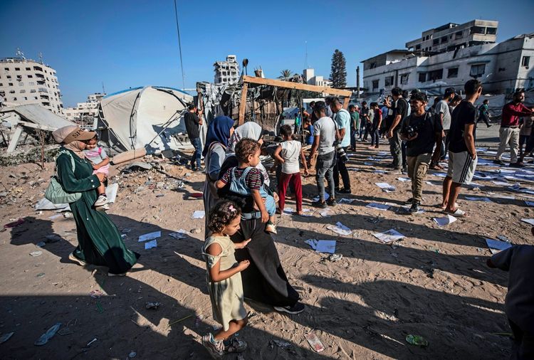 Eine Gruppe von Palästinensern inspiziert die Schäden nach einem israelischen Luftangriff in Gaza-Stadt am 27. August 2026. Im Vordergrund sind Erwachsene und Kinder auf einem von Trümmern und verstreuten Papieren bedeckten Gelände zu sehen. Im Hintergrund sind beschädigte Gebäude und Zelte erkennbar.