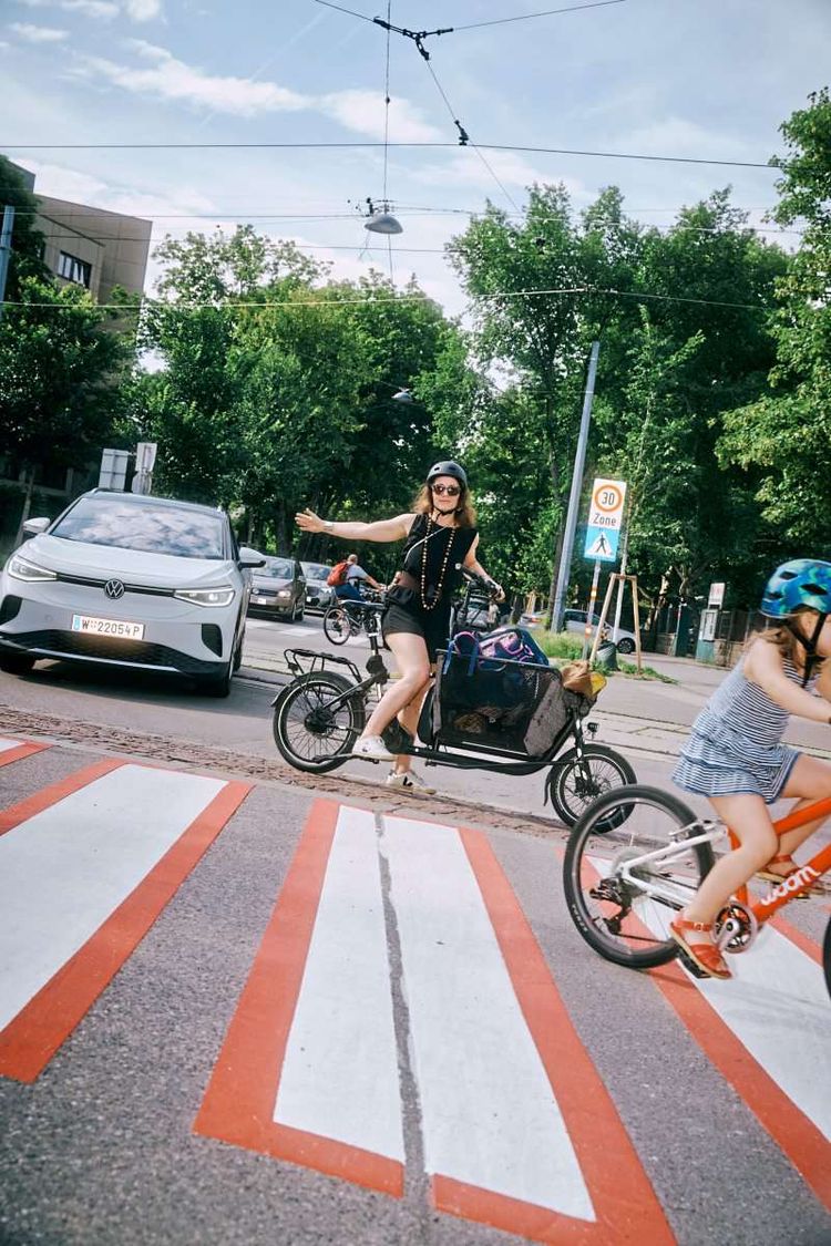 Elli steht mit dem Fahrrad auf der Straße und hält ein Auto auf, damit ihre Kinder überqueren können.