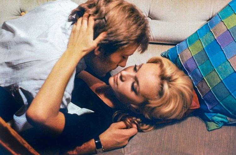 Ein Mann und eine Frau liegen auf einem Sofa. Der Mann beugt sich zu der Frau, während sie sich anschauen. Neben ihnen liegt ein farbenfrohes Kissen auf dem Sofa.