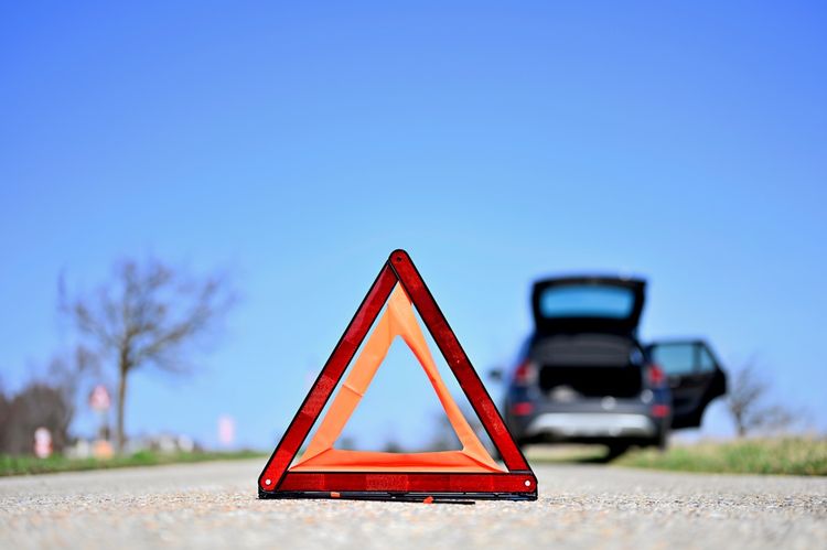 Ein Warndreieck steht auf einer Landstraße, im Hintergrund ist ein Auto mit geöffneter Heckklappe am Straßenrand zu sehen. Klarer blauer Himmel und unscharfe Bäume im Hintergrund.