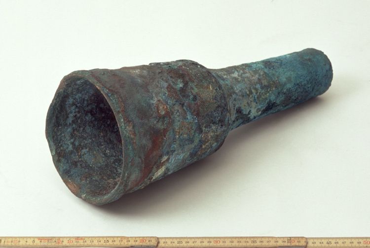 Eine verbeulte Kupferkanone, deren Form an ein Fernrohr erinnert. Ein beiliegendes Maßband zeigt etwa 50 cm Länge.