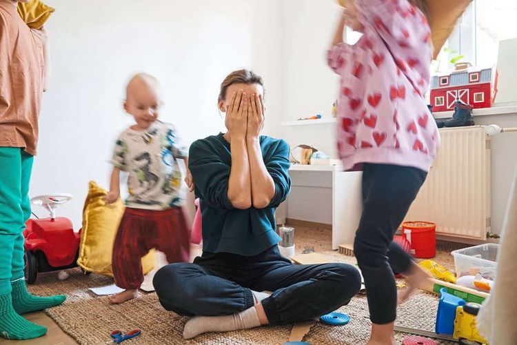 Eine Mutter sitzt auf dem Boden im Kinderzimmer und hält sich die Hände vors Gesicht, während drei Kinder um sie herumlaufen und toben