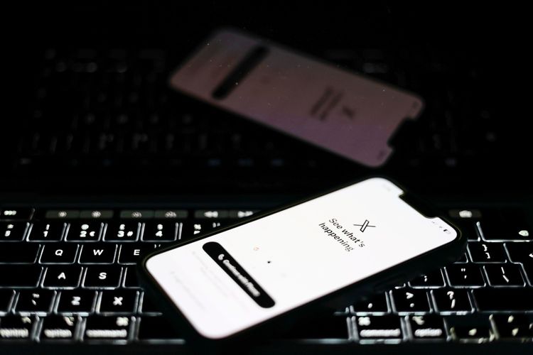 Ein Smartphone mit der geöffneten X-Anwendung (ehemals Twitter) liegt auf einer beleuchteten Laptop-Tastatur. Der Bildschirm zeigt die Startseite der App mit dem Text „See what's happening“. Im Hintergrund spiegelt sich das Smartphone-Display leicht auf der Laptop-Oberfläche. Die Szene ist dunkel gehalten, mit Fokus auf den Geräten.
