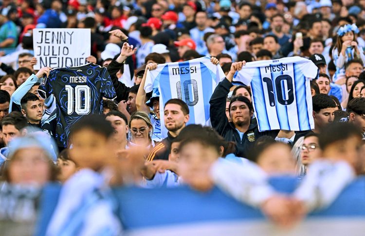 Argentinische Fans halten vor einem Spiel im Stadion verschiedene Lionel-Messi-Trikots hoch, darunter Heim- und Auswärtstrikots mit der Nummer 10. Im Hintergrund ist eine große Menschenmenge zu sehen.