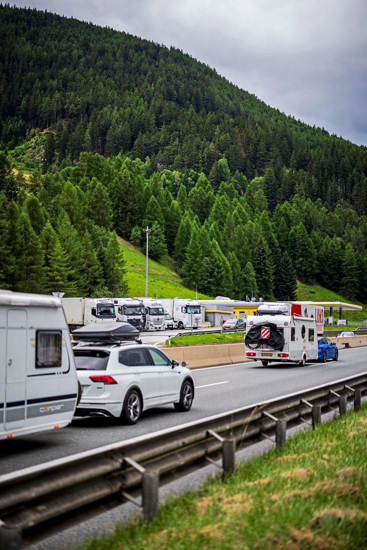 Ein Autobahnabschnitt am Brennerpass mit dichterem Verkehr, darunter Wohnwagen, Autos und Lkws. Im Hintergrund eine Rastanlage und bewaldete Berghänge. Bewölkter Himmel.