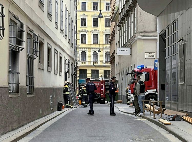 Straßenszene in der Wiener Innenstadt mit Einsatzkräften der Polizei und Feuerwehr nach einem teilweisen Gebäudeeinsturz. Feuerwehrfahrzeuge und Rettungspersonal sind vor Ort, während Rettungsmaßnahmen durchgeführt werden. Im Hintergrund sind Gebäude zu sehen, darunter eines mit dem Schriftzug 