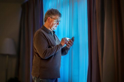 Ein Mann steht vor einem Fenster und blickt auf sein Smartphone.