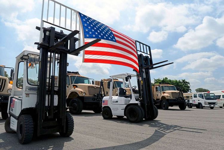 Zwei Gabelstapler, zwischen denen eine US-Fahne aufgespannt ist, dahinter Militärlastwagen.