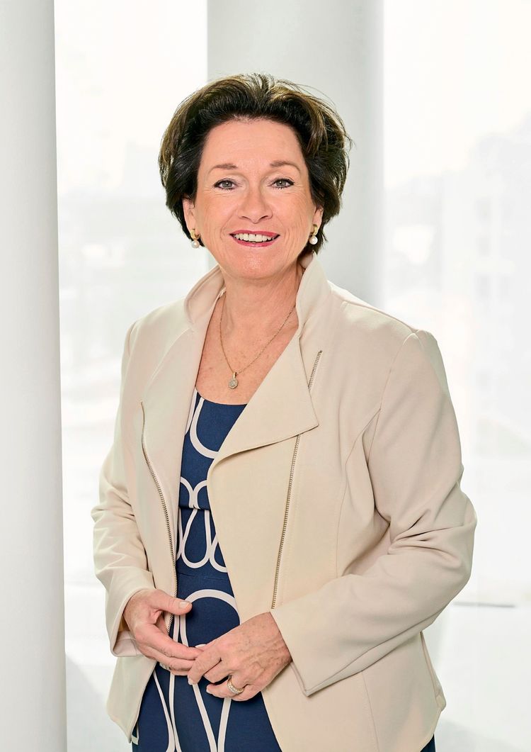 Eine Frau mittleren Alters mit schulterlangem dunklem Haar lächelt in die Kamera. Sie trägt einen cremefarbenen Blazer über einem dunkelblauen Kleid mit weißem Muster. Der Hintergrund ist hell und unscharf.