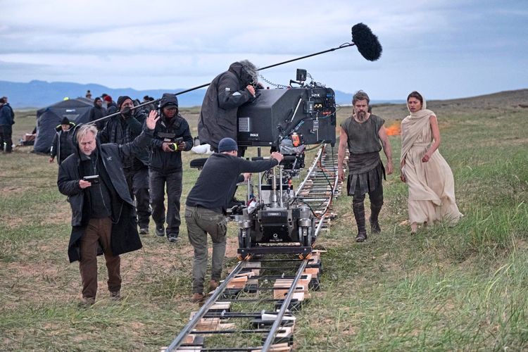 Eine Filmcrew dreht eine Szene in einer Graslandschaft. Links der Regisseur mit Drehbuch, in der Mitte eine Kamera auf Schienen mit Technikern, und rechts zwei Schauspieler in antiken Kostümen.