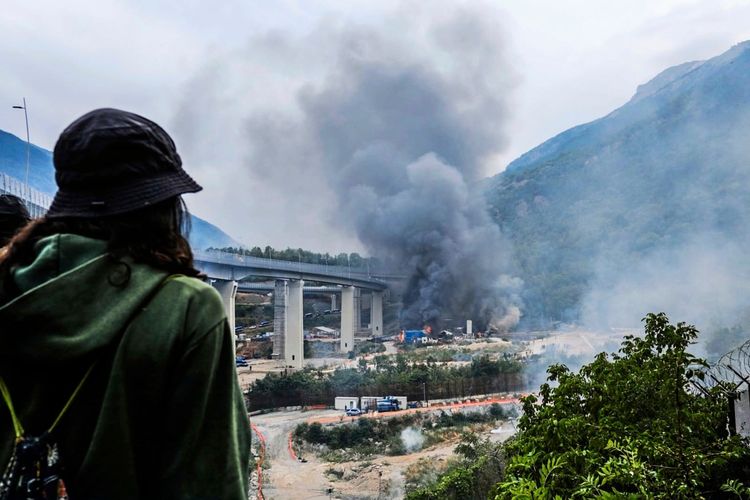 Eine Person mit grünem Hoodie und schwarzem Hut blickt auf eine Szene mit dichter schwarzer Rauchentwicklung nahe einer Baustelle. Im Hintergrund sieht man eine Brücke, Berge und Vegetation. Das Bild zeigt einen Protest mit Feuer und Rauch im Zusammenhang mit der umstrittenen Hochgeschwindigkeitszugstrecke Turin-Lyon (TAV) in Chiomonte, Susa-Tal, Norditalien.