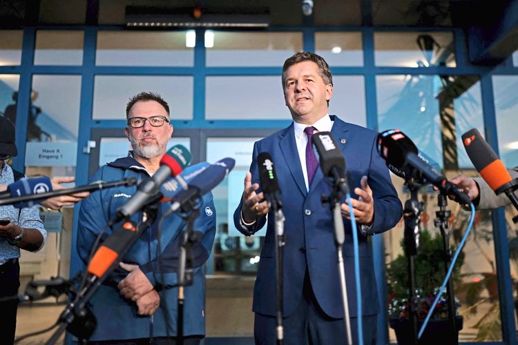 Zwei Männer stehen vor Mikrofonen und sprechen zu Pressevertretern. Der Mann rechts trägt einen dunklen Anzug mit Krawatte und gestikuliert mit den Händen, während der Mann links eine Brille und eine dunkelblaue Jacke trägt. Im Hintergrund ist ein Gebäudeeingang zu sehen.