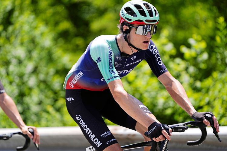 Ein Radfahrer des Decathlon CMA CGM Teams in einem Rennen, mit grünem Helm und mehrfarbigem Trikot, auf einer asphaltierten Straße mit grüner Naturkulisse im Hintergrund.