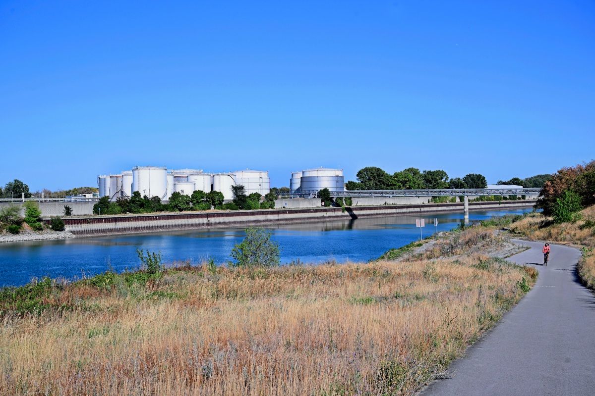 Industrieanlage mit mehreren großen silbernen Tanks in einer grünen Umgebung, die an einen Fluss grenzt. Im Vordergrund eine asphaltierte Fahrradstraße mit einer Person auf einem Fahrrad. Klarer blauer Himmel im Hintergrund.