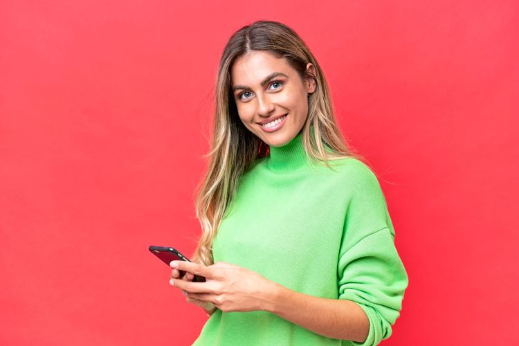 Junge Frau in einem grünen Pullover hält ein Smartphone in der Hand und lächelt vor einem roten Hintergrund.