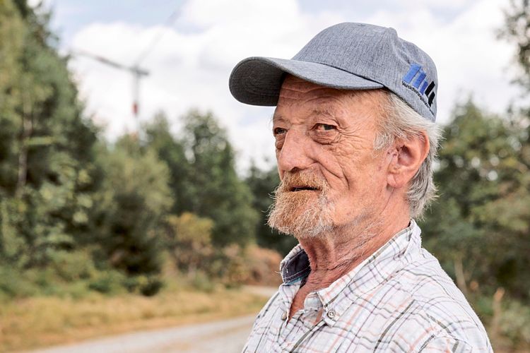 Ein älterer Mann mit grauem Bart und blauer Baseballkappe steht auf einem Schotterweg in einer ländlichen Umgebung. Im Hintergrund sind Bäume und ein Windrad zu erkennen. Der Himmel ist leicht bewölkt.