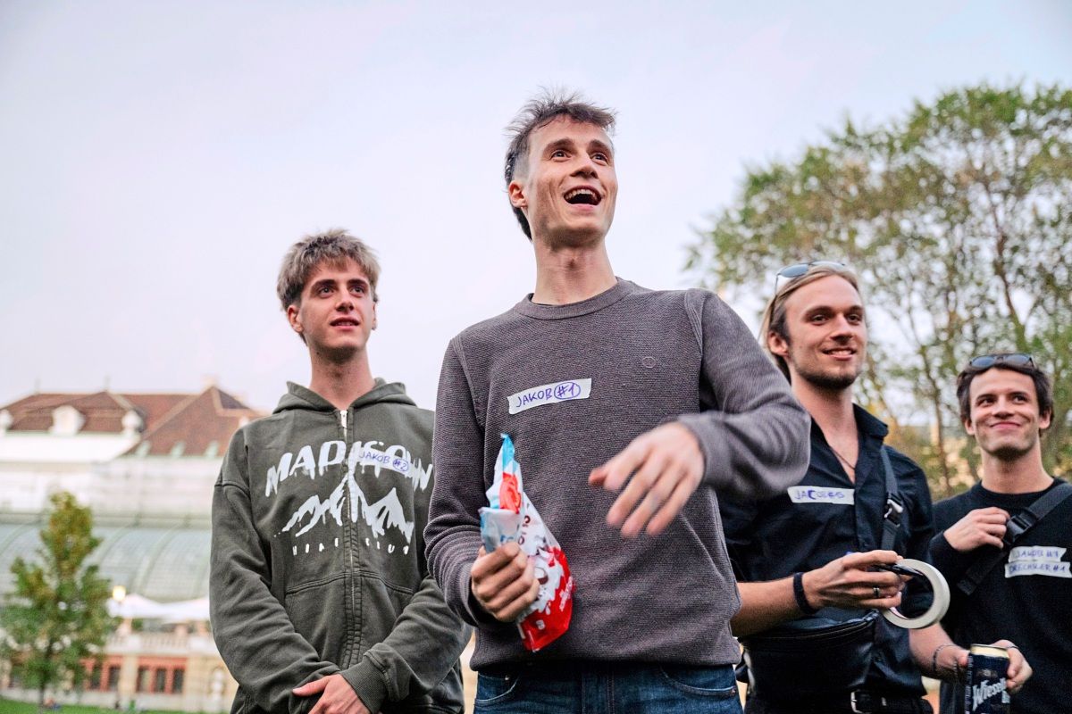 Eine Gruppe von vier jungen Männern steht im Freien und lacht. Zwei von ihnen tragen Namensschilder mit der Aufschrift „Jakob“. Im Hintergrund sind Bäume und ein Gebäude zu sehen, vermutlich bei Dämmerung. Einer der Männer hält ein kleines Geschenk.