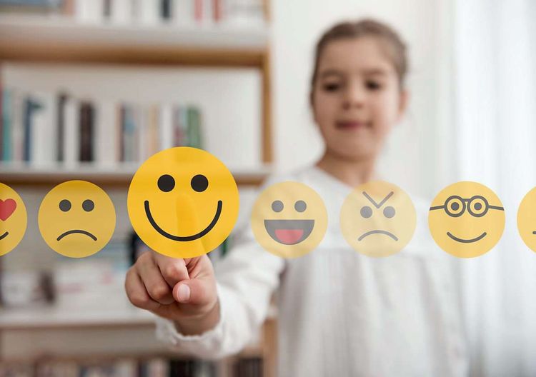 Ein Mädchen steht hinter einer Scheibe und tippt mit dem Zeigefinger auf einen lachenden Smiley, daneben hängen noch andere Emojis