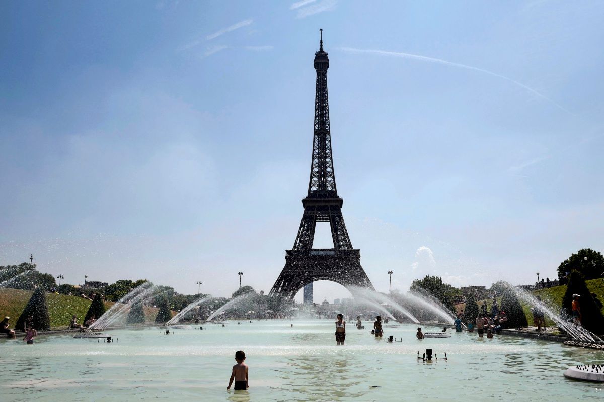 Menschen kühlen sich bei heißem Wetter in der Trocadero-Brunnenanlage vor dem Eiffelturm in Paris ab. Wasserfontänen sprühen, während Erwachsene und Kinder im flachen Wasser spielen. Klarer Himmel im Hintergrund.