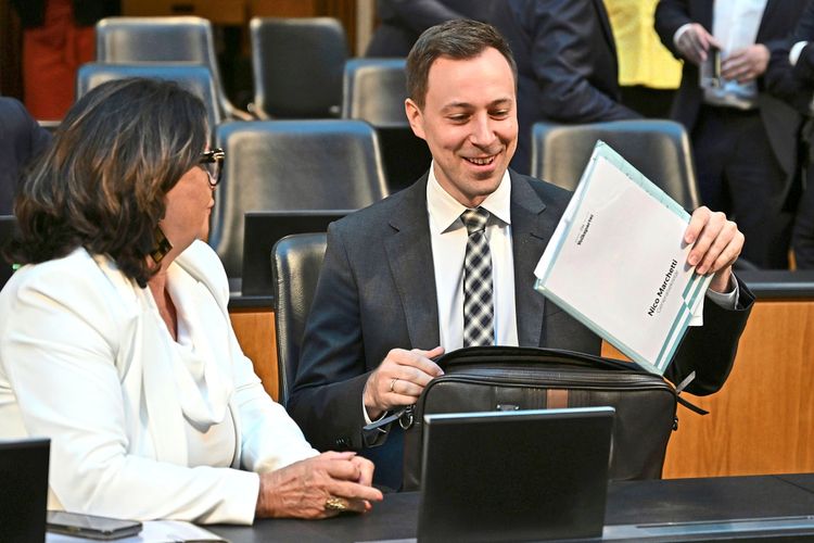 Zwei Personen sitzen in einem Sitzungssaal. Ein Mann im Anzug hält lächelnd eine Mappe mit der Aufschrift 