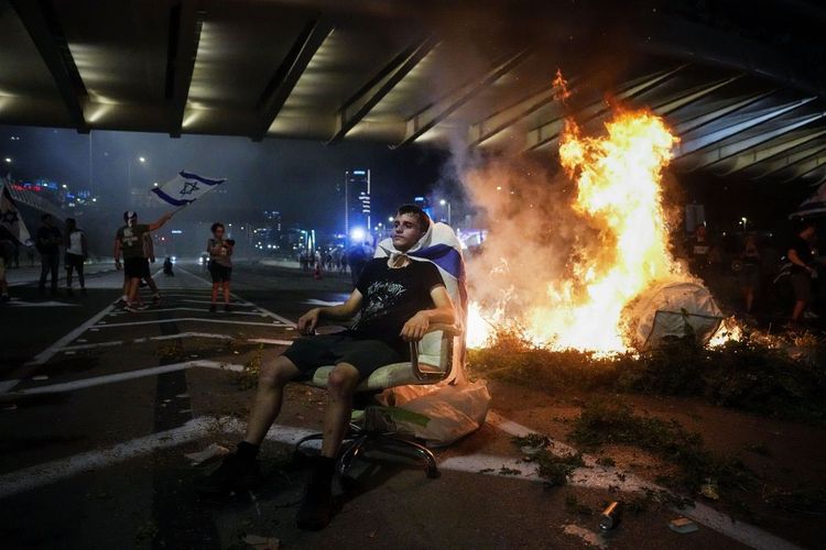 Ein Demonstrant sitzt auf einem Stuhl auf dem Ayalon Highway in Tel Aviv. Im Hintergrund ist ein Feuer zu sehen.