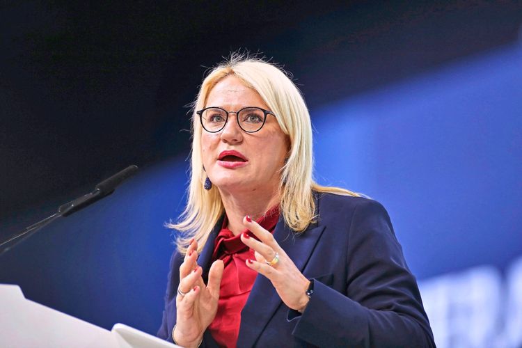 Eine Frau mit blonden Haaren und Brille spricht auf einer Bühne. Sie trägt einen dunkelblauen Blazer und ein rotes Oberteil. Hinter ihr ist ein blauer Hintergrund zu sehen.