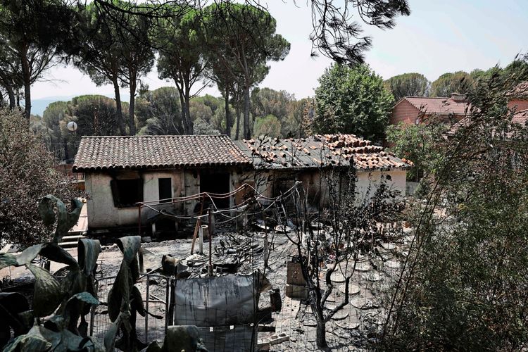 Ein ausgebranntes Haus steht im Vordergrund, beschädigt durch ein kürzliches Waldbrandereignis. Im Hintergrund sind Bäume und weitere Häuser sichtbar. Die Vegetation um das Haus herum ist ebenfalls stark verbrannt.