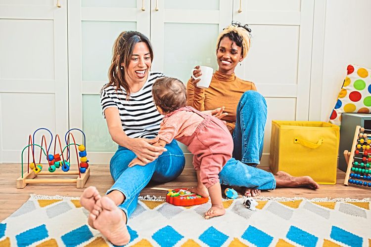 Zwei Frauen sitzen am Boden und spielen mit einem Baby, überall liegt Spielzeug