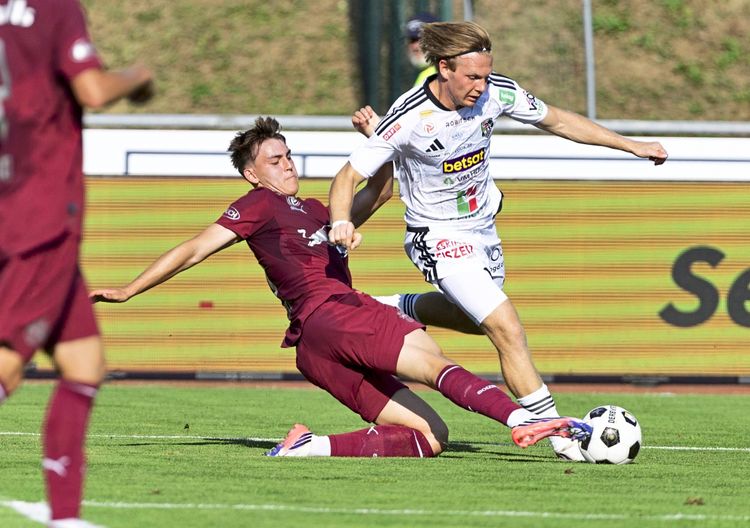 Zwei Fußballspieler im Zweikampf während eines Bundesliga-Spiels: Ein Spieler in weinrotem Trikot grätscht am Boden, während ein Spieler in weißem Trikot versucht, mit dem Ball nach vorne zu kommen. Das Spiel findet auf einem Fußballfeld statt, im Hintergrund ist eine Werbebande zu sehen.