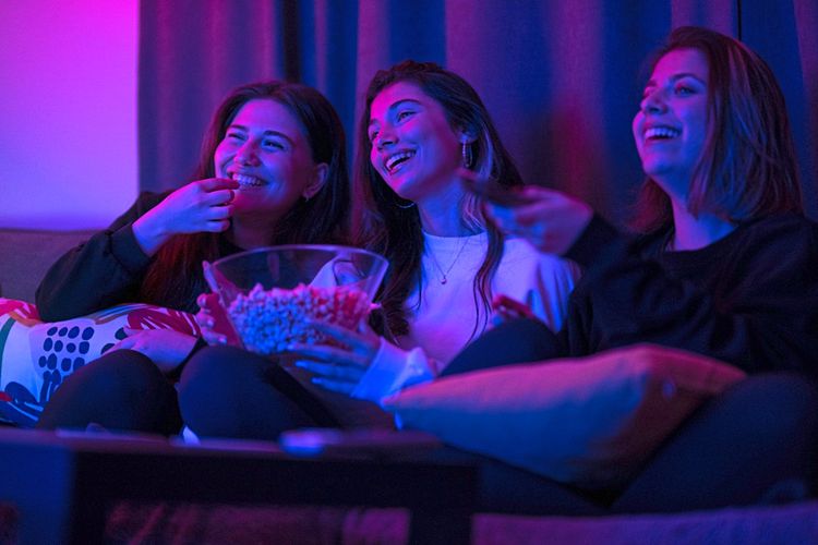 Drei Teenager-Mädchen sitzen Popcorn-essend und lachend beim Fernsehen auf einer Couch