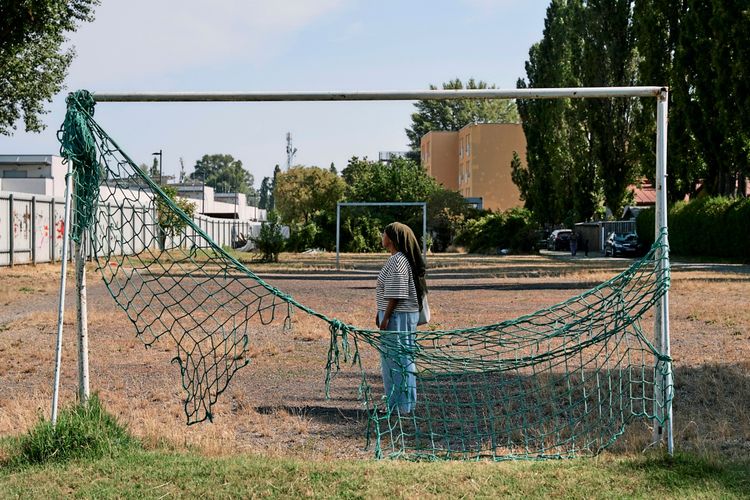 Die Autorin Ayaan Adan Mohamed steht in einem alten, beschädigten Fußballtor mit zerrissenem Netz auf einem trockenen, ungenutzten Feld in der Wohnsiedling Macondo, Zinnergasse 29. Im Hintergrund sind Gebäude, Bäume und geparkte Autos in einer städtischen Umgebung zu sehen.