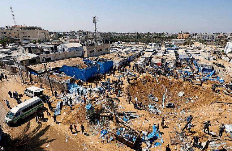 Luftaufnahme eines Geländes in Deir al-Balah im zentralen Gazastreifen mit einem großen Krater, der von einem nächtlichen israelischen Luftangriff verursacht wurde. Menschen durchsuchen die Trümmer um den Krater herum. Im Hintergrund sind Zelte zu sehen, die vertriebene Personen beherbergen.