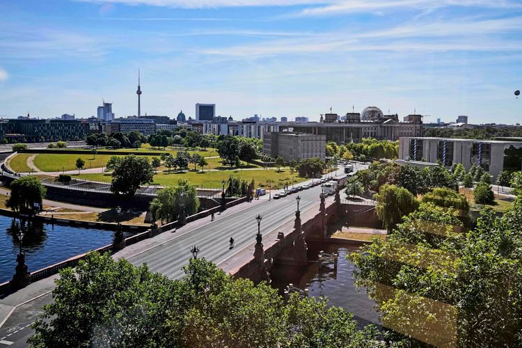 Blick aus dem neuen Übergangsbüro des deutschen Bundespräsidenten am 24. Juni 2026. Zu sehen sind die Moltkebrücke im Vordergrund, die Spree, das Bundeskanzleramt (rechts), das Reichstagsgebäude (zweite von rechts) und der Berliner Fernsehturm (links). Das Büro liegt gegenüber dem Bundeskanzleramt, während Schloss Bellevue renoviert wird. Im Vordergrund sind Grünflächen, Bäume und der Fluss erkennbar, unter strahlend blauem Himmel.